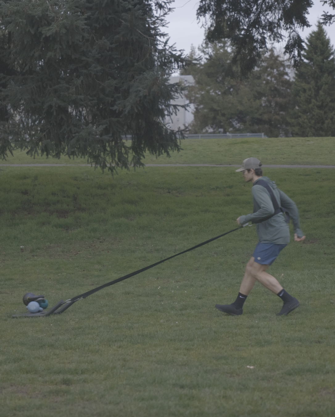 Connor pulling a weighted sled loaded with kettlebells across a park field using a shoulder harness