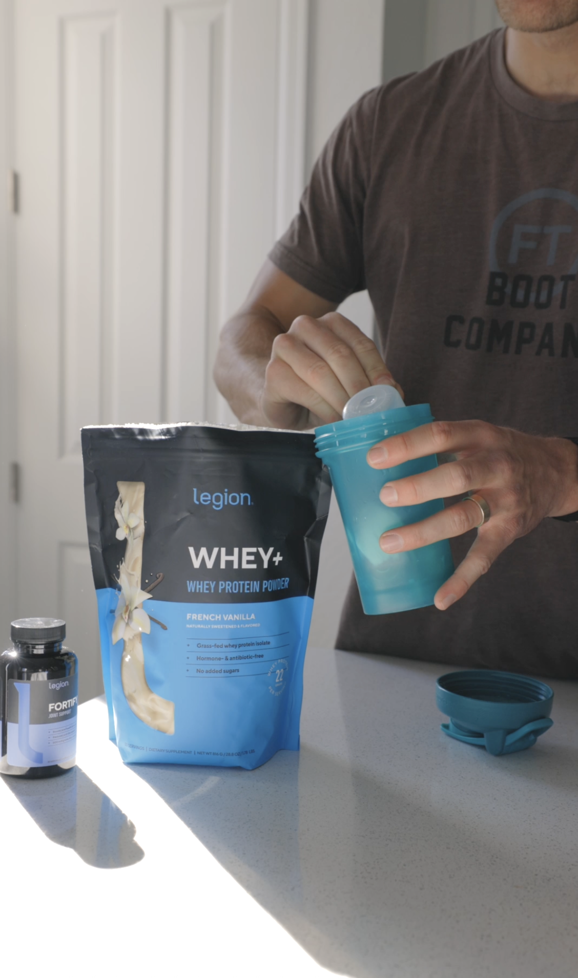 Scooping Legion Whey+ vanilla protein powder into a shaker bottle, with a bottle of Legion Fortify joint support on the counter