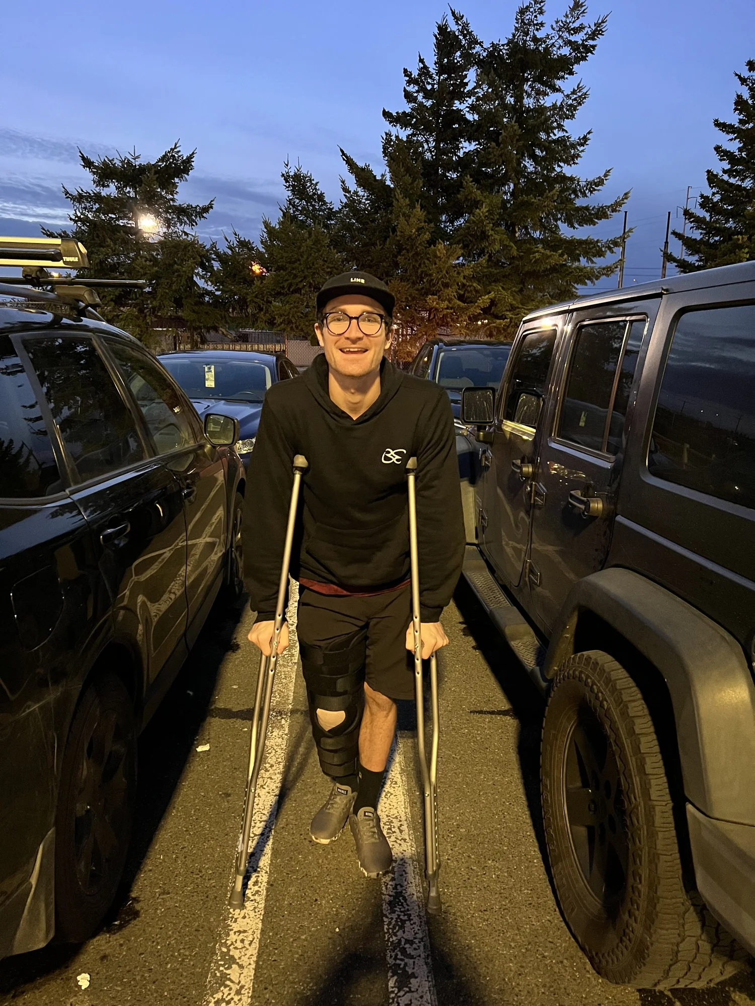 Connor on crutches in a parking lot at dusk, right leg in a full-length knee brace after ACL surgery, still smiling