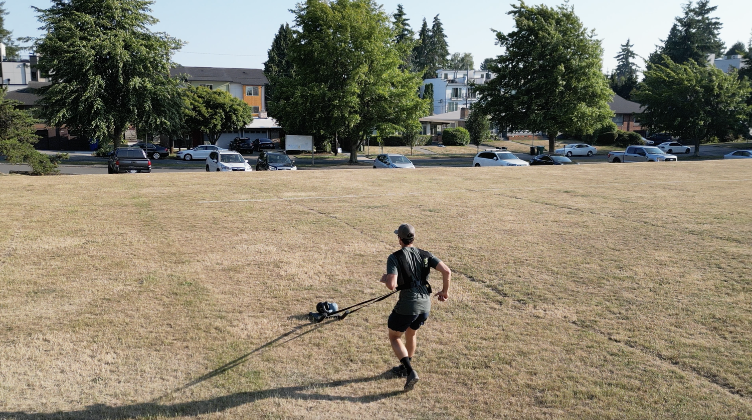 Connor dragging a weighted sled across a dry grass field with a harness