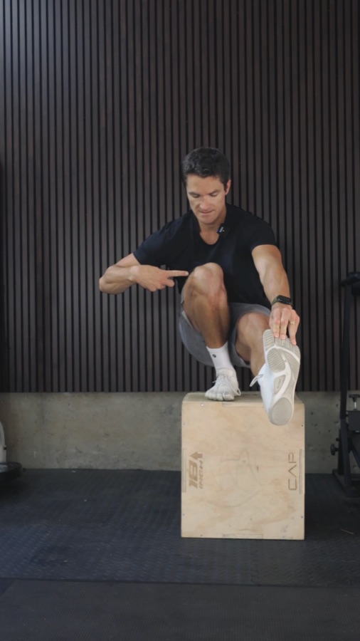 Connor balancing in a deep single-leg squat on a plyo box, pointing at his knee
